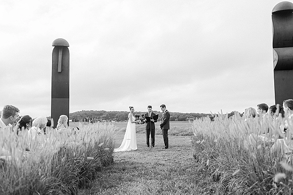Wedding ceremony in an open field with bride and groom exchanging vows before an officiant, guests seated nearby under wide sky horizon trees