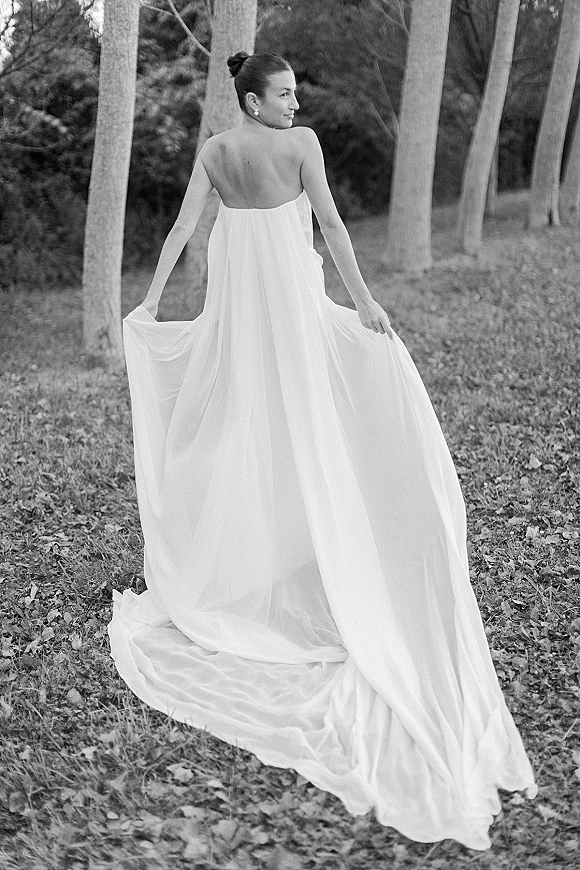 Bridal portrait in a strapless wedding dress with a long train, updo bun and earrings, looking back while holding the train in a leaf-strewn park.