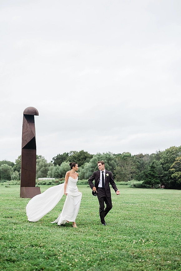 Couple portrait of bride and groom walking across a grassy field, bride lifting her long train in an overcast setting with a sculpture behind