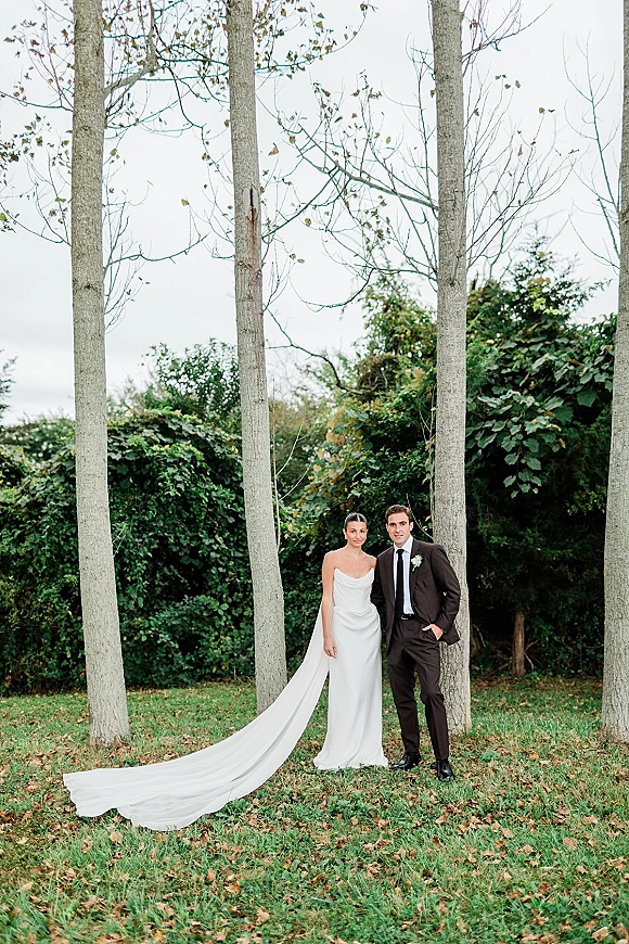 Couple portrait of bride in a strapless wedding dress with a long train and groom in suit on a leaf-strewn lawn under tall trees