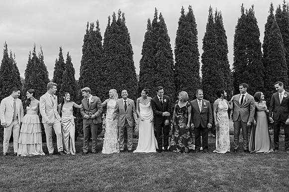 Wedding party portrait with bride, bridesmaids and groomsmen lined up in formal attire on a lawn with evergreen trees under a cloudy sky