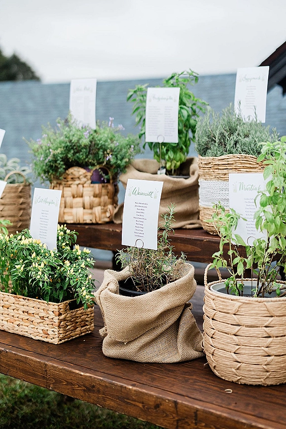 Escort card display with wedding escort cards tucked into potted herbs on wooden risers, burlap and woven baskets on an outdoor lawn table