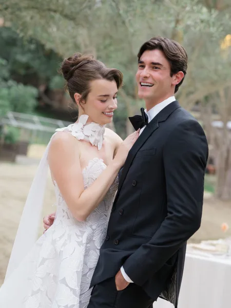 Couple portrait of bride holding groom’s lapel, wearing a lace high-neck gown and veil, beside groom in black tuxedo on a garden path