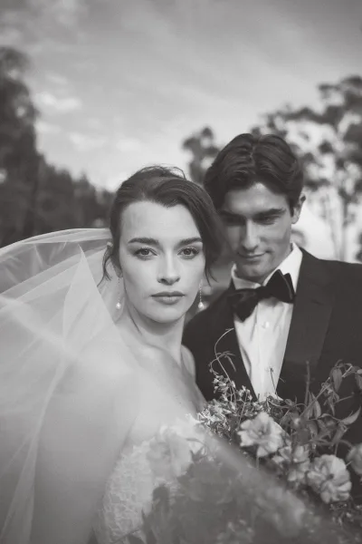 Couple portrait in a black and white wedding portrait style, bride in veil holding bouquet as groom in tuxedo stands behind her outdoors.