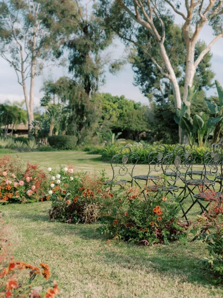 Ceremony seating with outdoor ceremony chairs in wrought iron lined along a garden lawn aisle bordered by coral ground florals and greenery