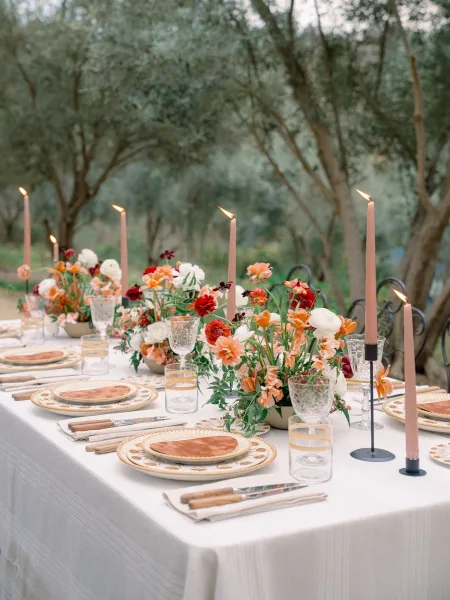 Reception tablescape with floral centerpieces and pink taper candles on a long outdoor reception table, set under trees in a garden setting