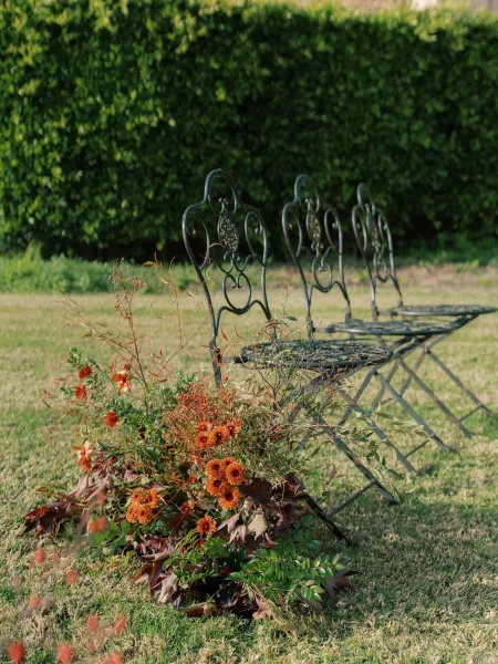 Ceremony aisle decor with an aisle end floral arrangement of burnt orange flowers and greenery beside a wrought iron bench on a grass lawn