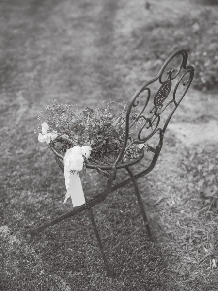 Wedding bouquet with white ribbon resting on a wrought iron chair, surrounded by garden foliage and grass lawn, hand-tied greenery detail