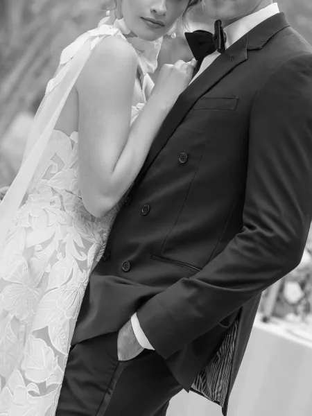 Couple portrait in a black and white wedding portrait, bride and groom close up embracing, her veil draped over his tuxedo in a blurred outdoor setting