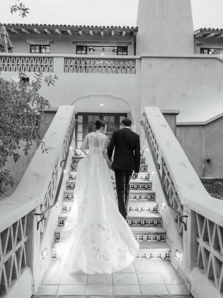 Newlywed exit as bride and groom walk away holding hands up a tile staircase, her dress train trailing beside his tuxedo in black and white