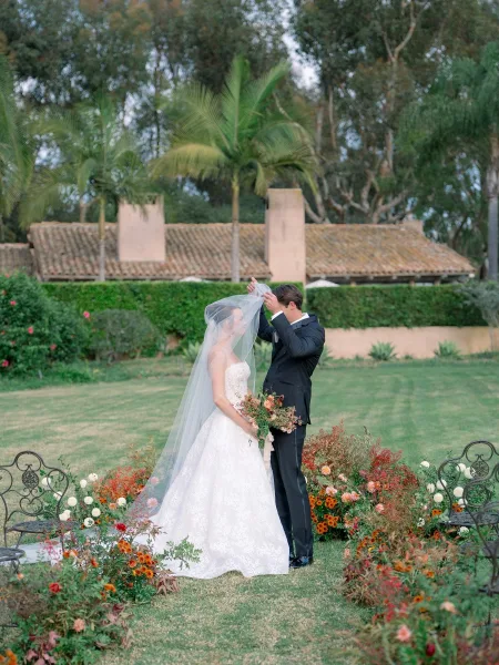 First look moment as the groom lifting veil over the bride’s face, bouquet in hand on a garden lawn with palm trees behind