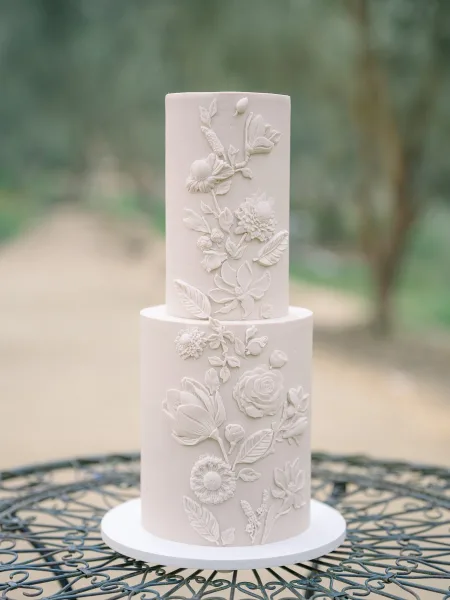 Wedding cake with white buttercream and raised floral appliques on a cake board atop a wrought iron table in an outdoor garden setting