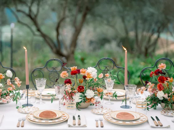 Reception tablescape with floral centerpieces and blush taper candles on white linens, patterned plates and glassware set outdoors amid garden greenery