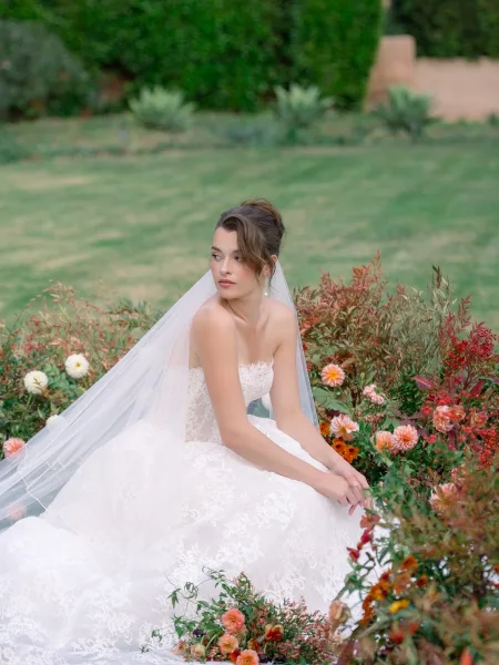 Bridal portrait of a bride seated among garden flowers in a strapless lace wedding dress and cathedral veil, looking over her shoulder outdoors