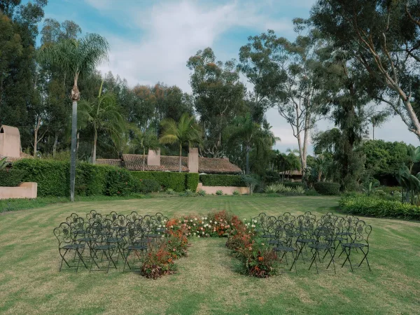 Ceremony setup with outdoor wedding ceremony seating in a semicircle of wrought iron chairs, low orange and white aisle flowers on a garden lawn
