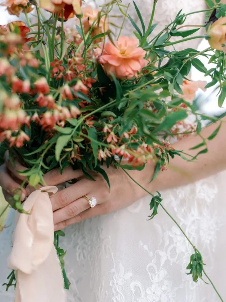 Bridal bouquet with peach wedding bouquet blooms and greenery, tied with ribbon, held by a bride showing an oval diamond ring on lace dress