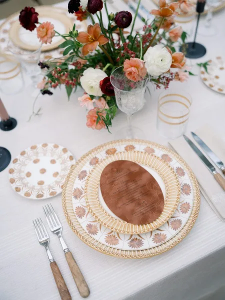 Reception tablescape with a floral centerpiece, patterned plates and menu card, black taper candles, and glassware on a white linen table