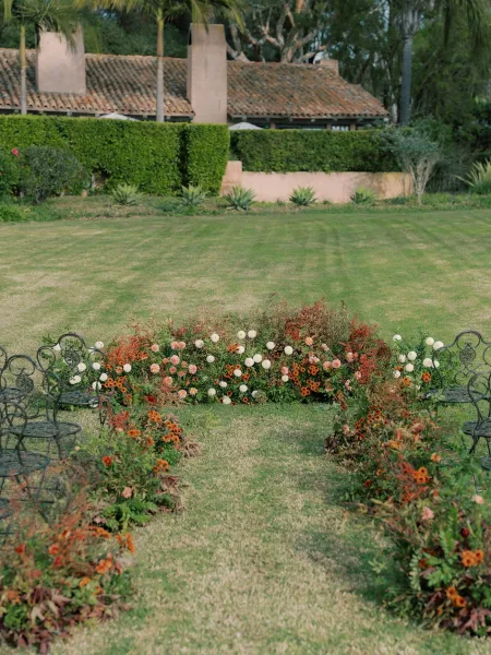 Ceremony aisle decor with outdoor ceremony aisle flowers and lush greenery beside wrought iron chairs on a lawn with palms and stucco building