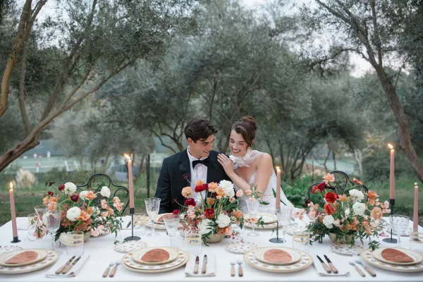 Couple portrait of bride and groom at table, her sheer neck scarf beside pink taper candles and florals in a garden setting