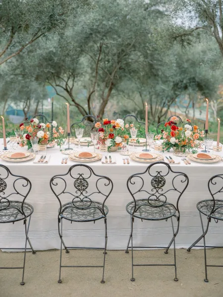 Reception tablescape with white linen table setting, floral centerpieces, and tapered candles on a long banquet table outdoors among trees