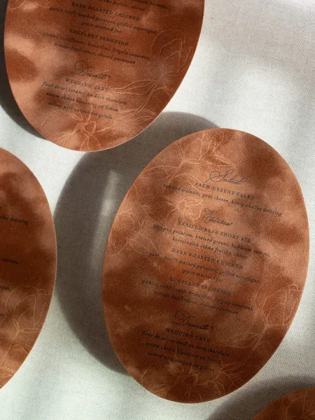 Wedding menu cards in an oval wedding menu style on white linen, printed on brown paper with delicate floral line art and text