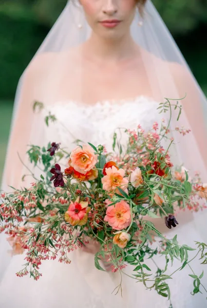 Bridal bouquet of peach wedding bouquet flowers and greenery held against a lace wedding dress and veil, framed by garden foliage