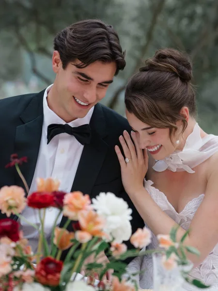 Couple portrait of bride and groom laughing as she shows her engagement ring, leaning together in greenery, with pearl earrings accent