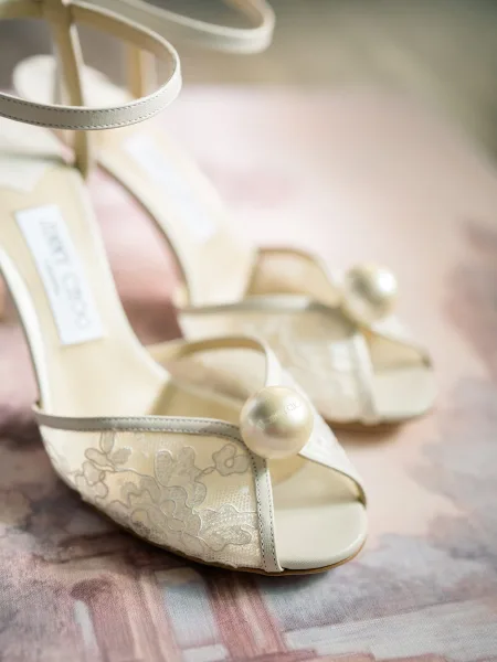Bridal shoes in a wedding heel close up, ivory ankle-strap heels with lace detailing and pearl embellishments on a pink surface