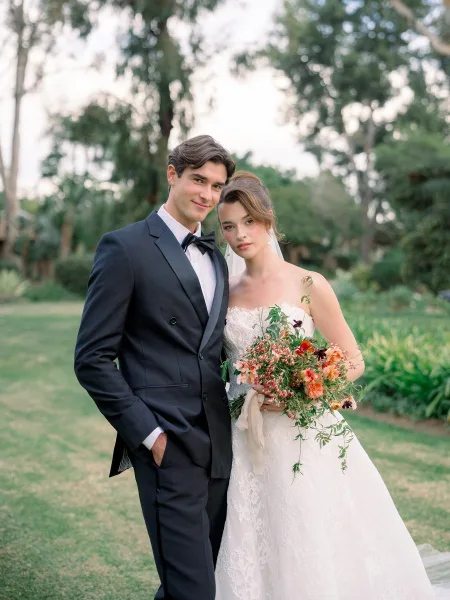 Couple portrait of bride and groom posing on a garden lawn, bride in veil holding bouquet beside groom in black tuxedo and bow tie