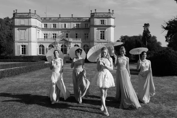 Bride and bridesmaids lead a bridal party walking with parasols across a sunlit garden lawn before a grand mansion facade.