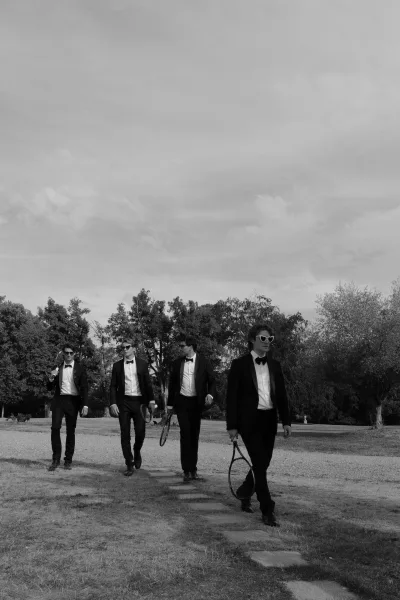 Groomsmen portrait becomes a groomsmen walking photo as they stride, playfully carrying tennis rackets across a stone path.