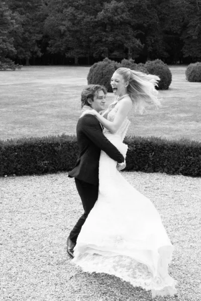 Couple portrait with groom lifting bride in a fitted bridal gown, laughing as they spin on a garden lawn bordered by hedges and trees.