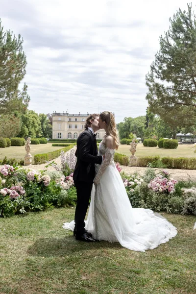 Wedding kiss portrait of a bride and groom in a garden, her long train brushing a gravel path by hedges and a distant historic building.
