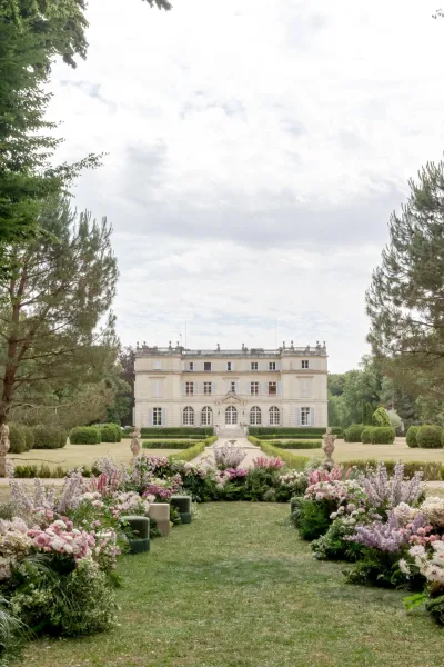 Ceremony aisle design featuring lush florals showcases refined garden ceremony decor before a stately mansion facade and formal hedges.