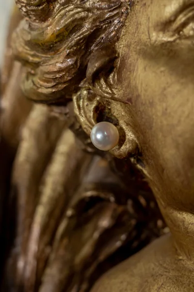 Pearl stud earring, a single pearl earring, rests against a gold sculpture on a textured gold metallic background.
