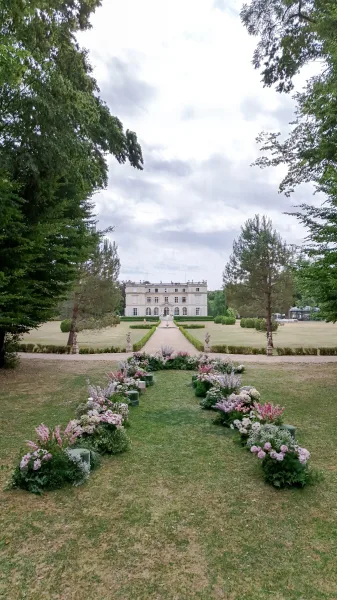 Ceremony aisle design with low florals lining a gravel path in an outdoor ceremony setup on a lawn before a mansion and fountain.