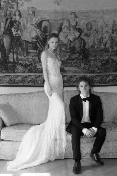 Couple portrait in a black and white wedding photo, bride in strapless lace gown with a pearl necklace beside a seated groom on an upholstered sofa.