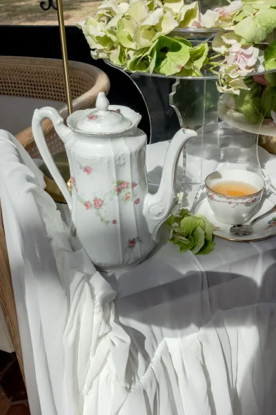 Tea set detail with a porcelain teapot and hydrangeas on a tablecloth by a window, with a wicker chair and lawn visible beyond.
