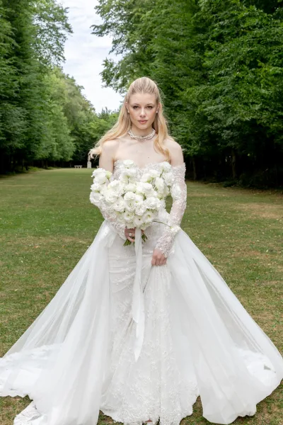 Bridal portrait of a bride holding a white bouquet in an off-the-shoulder lace gown, standing on a tree-lined garden path beneath cloudy skies.