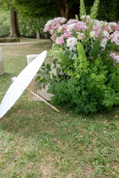 Wedding floral arrangement styled as outdoor wedding flowers, shaded by a white paper parasol on a grassy lawn framed by trees.