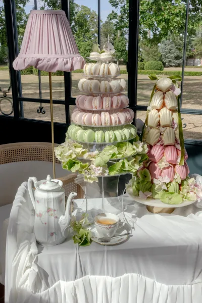 Macaron tower anchors a wedding dessert table, paired with a madeleine tower, set by glass windows framing garden trees and lawn.