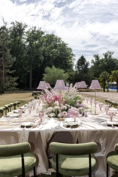 Reception tablescape on an outdoor reception table with pink glassware, styled beneath garden trees on a green lawn under open sky.