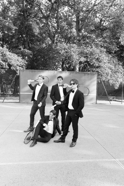 Groomsmen portrait that inspires groomsmen photo ideas, with the group posing with tennis rackets on a tennis court by a practice wall and fence.