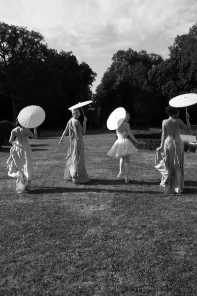 Bridal party photo of bridesmaids with paper parasols strolling across a grass lawn, framed by garden trees and hedges in soft summer light.