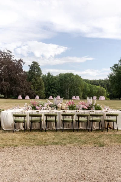Reception tablescape for an outdoor reception table, styled with table lamps, set on a garden lawn bordered by trees and a gravel path.