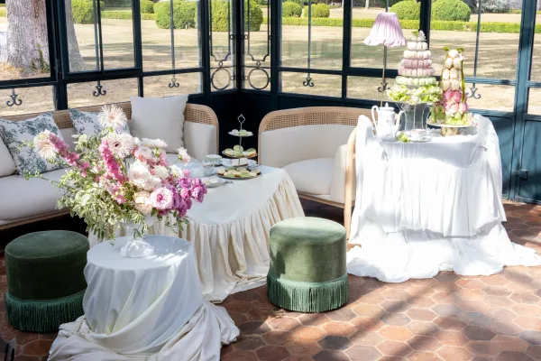 Wedding lounge setup with reception lounge seating centered on a rattan sofa, arranged beneath airy conservatory windows overlooking a garden.