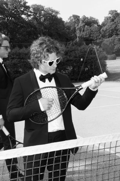 Groom portrait of a groom in tuxedo holding a tennis racket on a tennis court by the net, with trees and a chain-link fence behind.