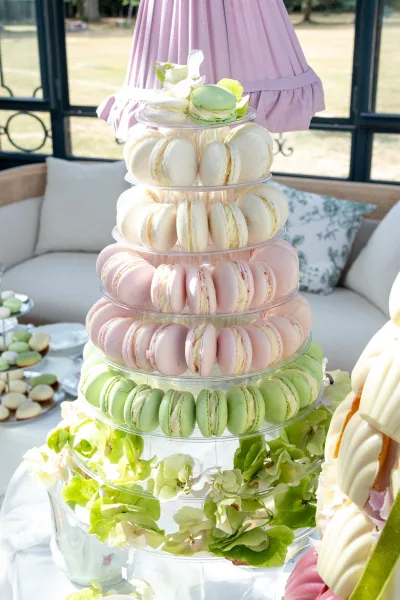 Macaron tower, a French macaron display on an acrylic tiered stand, styled beside teacups with a sofa and garden lawn beyond.