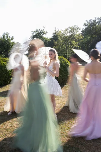 Bridal portrait of a bride with bridesmaids beneath paper parasols in a sunlit garden, with clipped hedges and a bright sky around them.