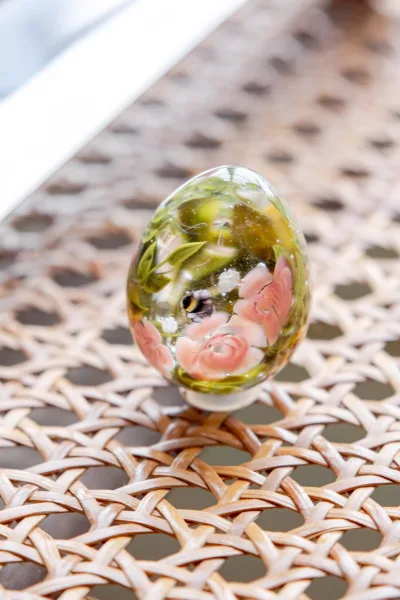 Glass paperweight shaped like a floral glass egg, encasing delicate roses, resting on a woven rattan background.